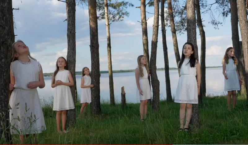 dziewczynki w białysch sukienkach stoją wśród drzew. W tle widać zalew.