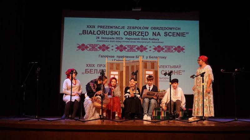 na scenie siedzi młodzież w białorsukich strojach. W tle wyświetlany jest plakat z wydarzenia