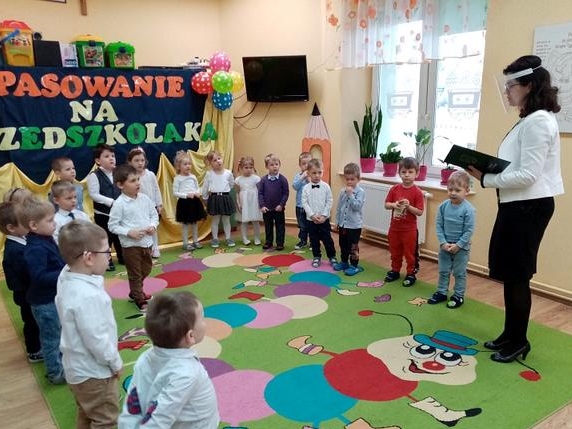 Na zdjęciu przedszkolaki stoja ustawione wokół krawędzi kolorowego dywanu. Po prawej stronie stoi pani dyrektor, w ręku trzyma książkę.