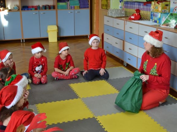 Zdjęcie przedstawia dzieci i panią siedzących na żółto-szarym dywanie. Wszyscy maja na sobie czerwone elementy garderoby. Pani trzyma przed soba zielony worek.