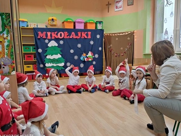 Na zdjęciu dzieci siedzą na podłodze, ubrane są w kolorach biało - czerwonych. Po prawej stronie siedzi kobieta. Za dziećmi widać czerwony napis: Mikołajki.