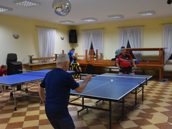 Na zdjęciu widac dwa stoły do tenisa stołowego. Zawodnicy ustawieni po przeciwnych stronach stołów, w ręce trzymają paletkę do gry, odbijając piłeczkę.