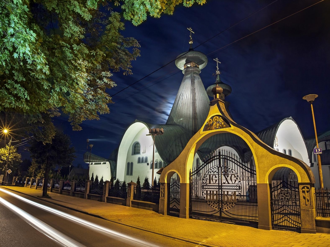 zdjęcie przedstawia świątynię w nocnej aurze, widoczna jest brama oraz przednia część cerkwi wraz z główną kopułą; w lewym górnym rogu znajduje się zielona korona drzewa 