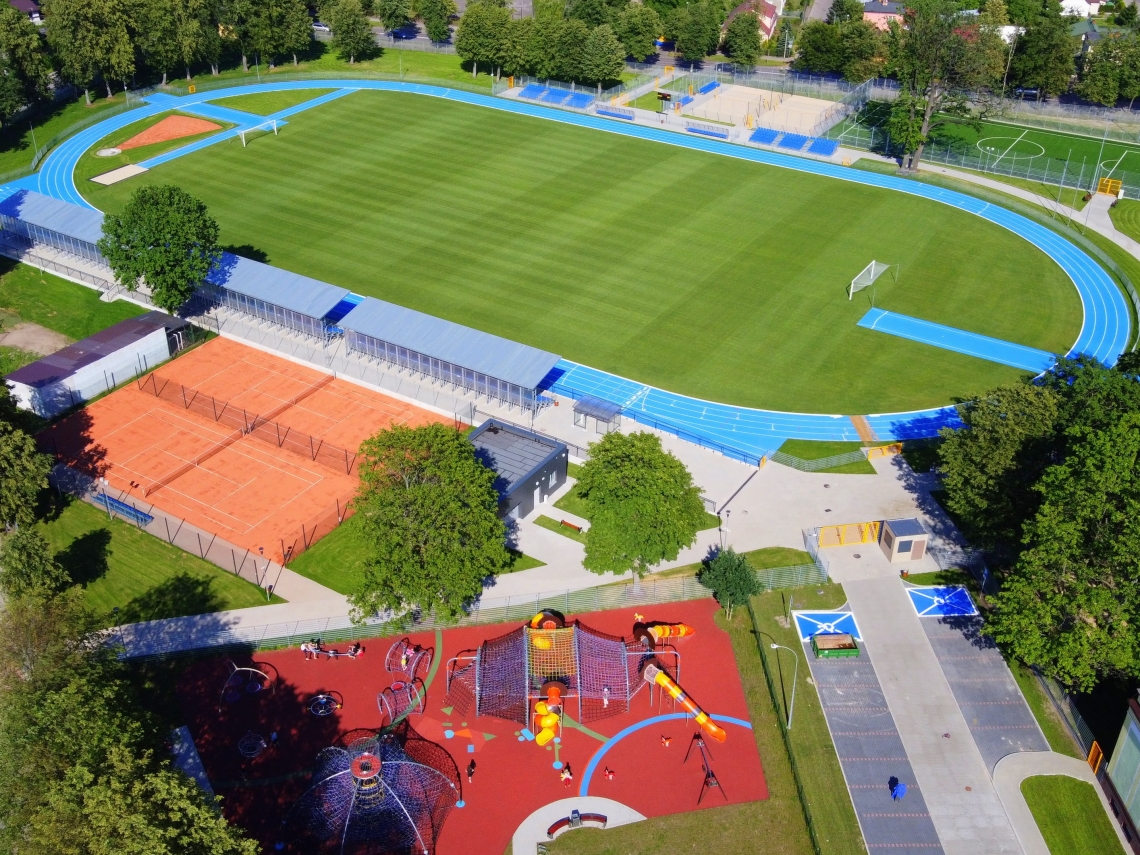 teren Ośrodka Sportu i Rekreacji z lotu ptaka; w centralnym punkcie boisko sportowe pokryte zieloną murawą, wokół niego wyznaczona jest niebieska trasa do biegów; z prawej strony widoczna jest Rodzinna Strefa Zabaw oraz kort tenisowy