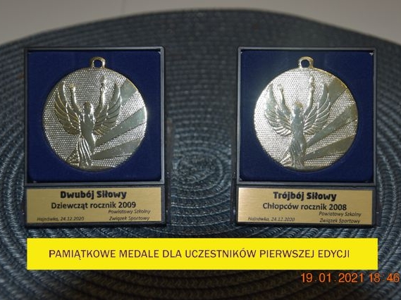 Na zdjęciu na szarej podkładce leżą dwa granatowe pudełka z medalami w kolorze srebrnym. Pod spodem na żótym tle napis: Pamiątkowe medale dla uczestników pierwszej edycji.