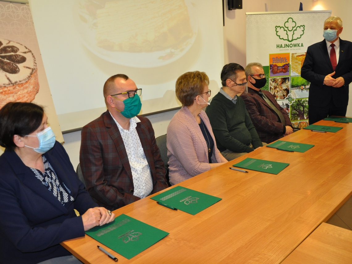 Na zdjęciu pięć osób siedzi za stołem, przed nimi zielone teczki. Po prawej stronie stoi Burmistrz.