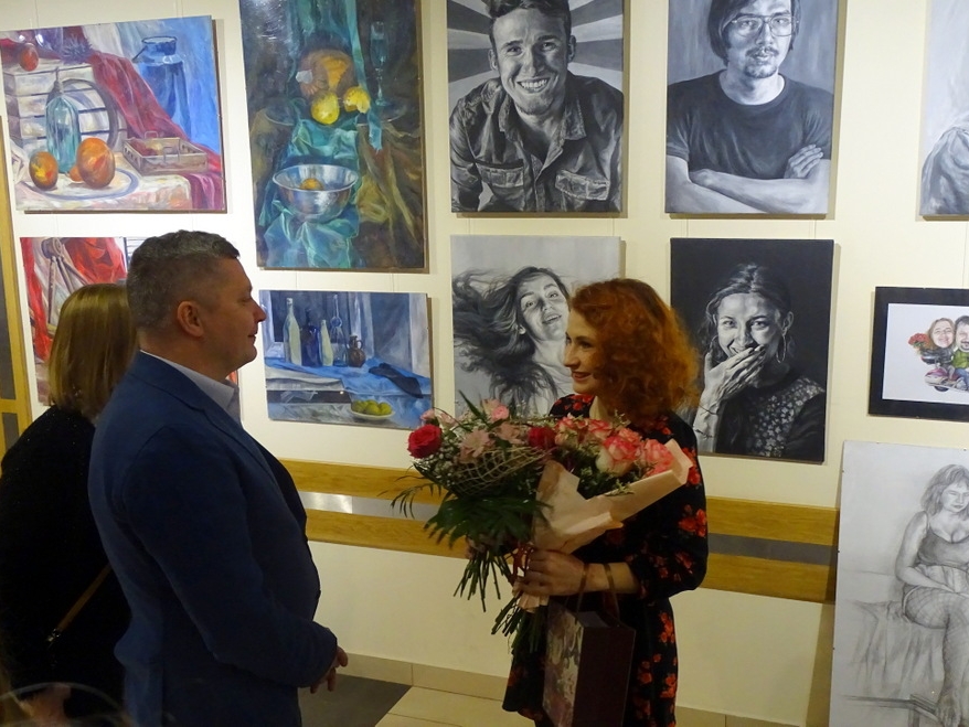 autorka wystawy stoi z kwiatami przed ekspozycją swoich prac. Dwie ososby gratulują jej udanej wystawy.