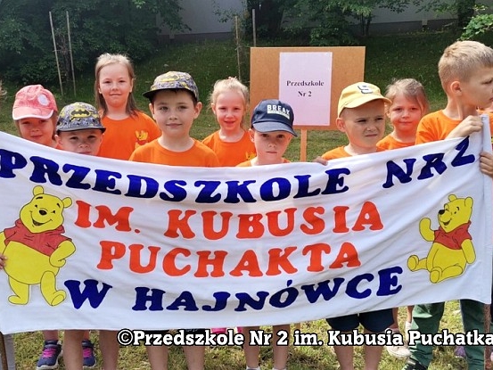 Dzieci w pomarańczowych koszulkach trzymają transparent z nazwa przedszkola, do którego uczęszczają