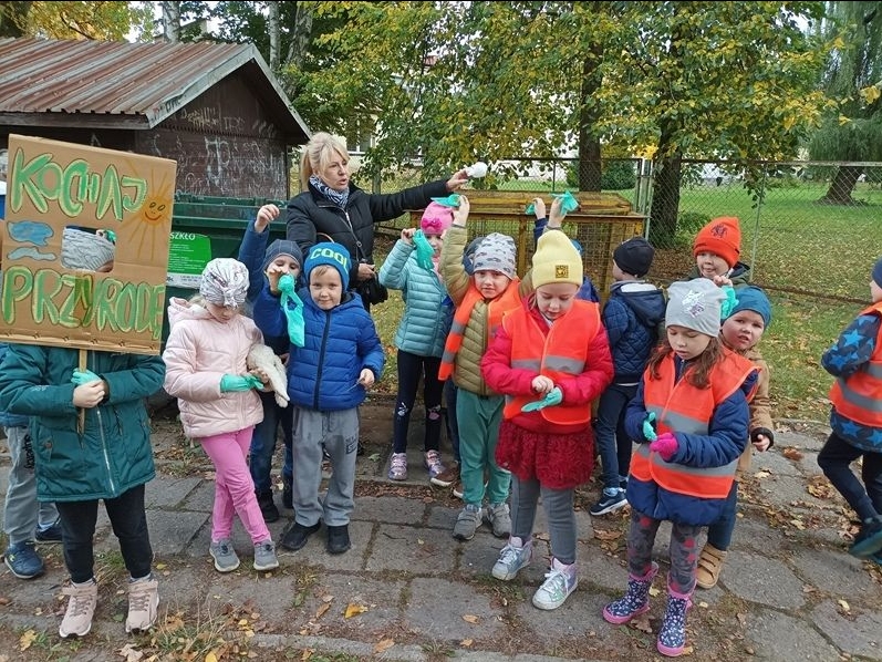 Dzieci stoją przed kontenerem i pozują wraz z nauczycielką do zdjęcia, trzymają w rękach gumowe rękawiczki i transparent z napisem: Kochaj Przyrodę!