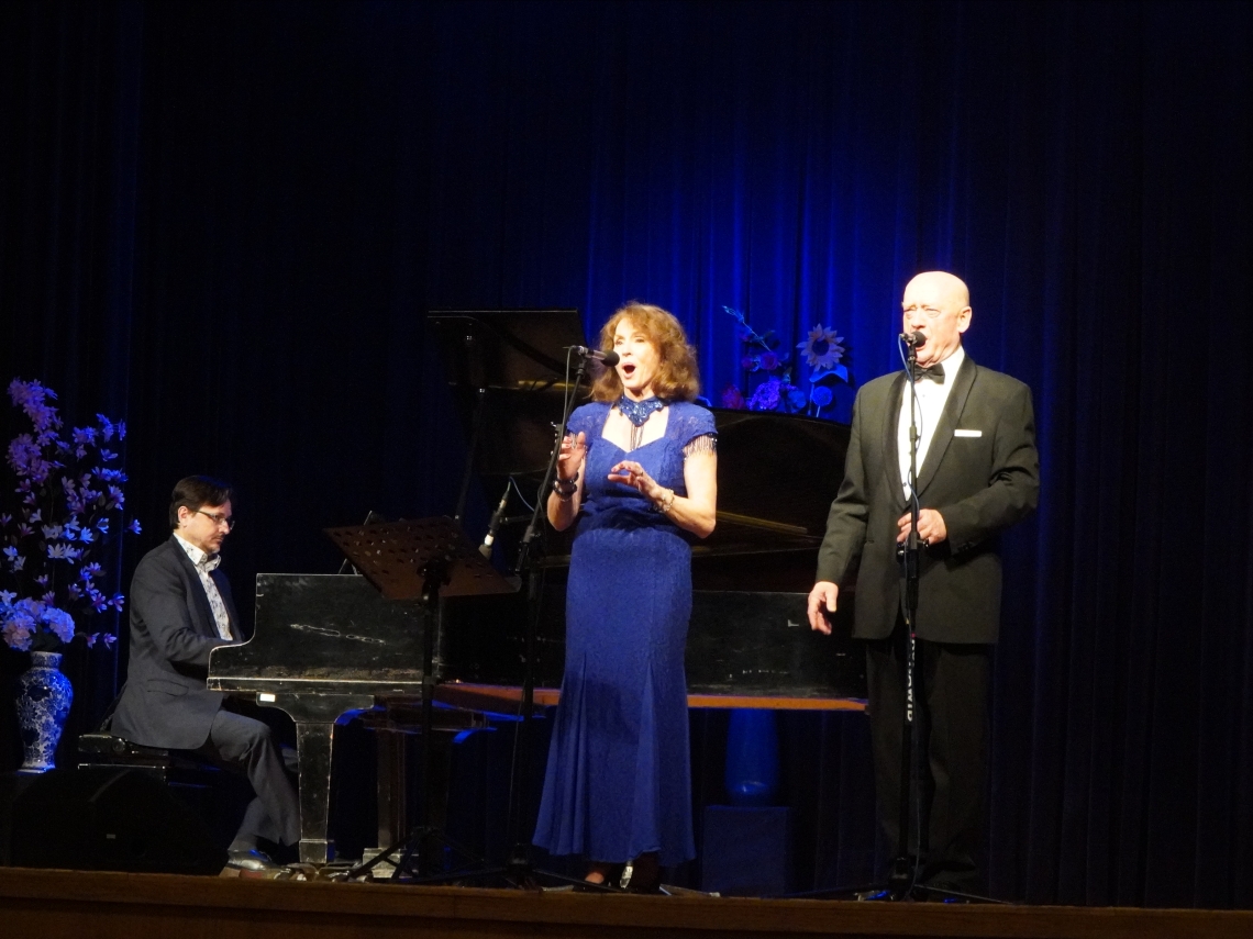 Kobieta i mężczyzna ubrani na galowo stoją na scenie. Obok siedzi przy fortepianie mężczyzna