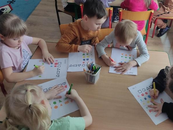 Dzieci siedzą przy stolikach i kolorują figury.