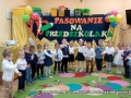 Dzieci stoją ustawione wokół kolorowego dywanu. Za nim napis na ścianie: Pasowanie na przedszkolaka.