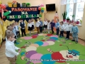 Dzieci ubrane w galowe stroje stoją ustawione wokół kolorowego dywanu.