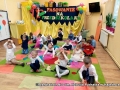 Dzieci siedzą na dywanie, trzymają ręce nad głowani tworząc z nich wzór czapek. Za nimi na ściane napis: Pasowanie na przedszkolaka.