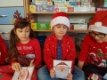 Na zdjęciu troje dzieci siedzi i trzyma rysunki świętego Mikołaja. Za nimi na półkach leżą gry planszowe.