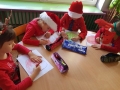Na zdjęciu dzieci siedzą przy stoliku i rysują kredkami na kartkach czerwone worki świętego Mikołaja. Na stolikach leża równiez piórniki na kredki.