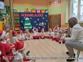 Na zdjęciu dzieci siedzą na podłodze, ubrane są w kolorach biało - czerwonych. Po prawej stronie siedzi kobieta. Za dziećmi widać czerwony napis: Mikołajki.