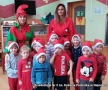 Zdjęcie grupowe. Dzieci mają na głowach czapki mikołaja, za nimi stoja dwie kobiety. Jedna z nich jest przebrana za elfa, ma na sobie czerwono - zielony strój i czerwona czapkę.