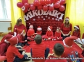 Na zdjęcie dzieci z paniami siedzą na czerwonym kocu, za nimi na czerwonym tle biały napis: Mikołajki.
