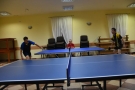 Na zdjęciu widac dwa stoły do tenisa stołowego. Zawodnicy ustawieni po przeciwnych stronach stołów, w ręce trzymają paletkę do gry, odbijając piłeczkę.