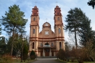 frony budynku kościoła wykonany z pomarańczowej cegły; wyróżniaa go dwie strzeliste wieże po obu stronach; do kościoła prowadzi ścieżka wyłożona kostką; obok budynku rosną wysokie sosny