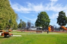 zdjęcie przedstawia widok na plac zabaw - w oddali widać urządzenia rekreacyjne, wiatę z ławeczką solarną, drewniane leżaki; wokół rosnie zielony trawnik