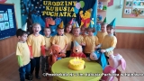 Zdjęcie pokazuje dzieci z grupy Tygryski ubrane w czerwone koszulki i urodzinowe czapeczki, które siedzą przy stole spożywając poczęstunek na tle dekoracji.