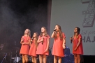 pięć dziewczynek w różowych sukienach wykonuje na scenie piosenkę