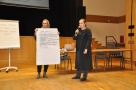 Uczestnicy prezentują wyniki swoich prac, na zdjęciu dwie osoby, jedna trzyma dużą kartkę, druga mówi do mikrofonu