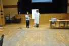 Uczestnicy prezentują wyniki swoich prac, na zdjęciu dwie osoby, jedna trzyma dużą kartkę, druga mówi do mikrofonu