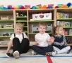 Troje dzieci siedzi na podłodze, za nimi stoją regały z zabawkami.