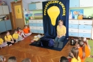 Dzieci i pani ubrani w żółte koszulki siedzą na dywanie, nad panią ogromna żarówka i napis "Oszczędzamy".