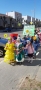 Dzieci spacerują po mieście, z przodu kolumny idą dziewczynki ubrane w kolorowe suknie.