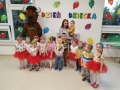 Grupa dzieci z nauczycielką stoją przed napisem "dzień dziecka", dziewczynki ubrane sa w czerwoje tiulowe spódniczki.