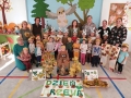 Zdjęcie przedstawia grupę dzieci z nauczycielkami oraz panią leśnik podczas pamiątkowego ustawienia z plakatami drzew z okazji Dnia Drzewa.