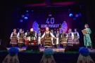 Na scenie stoją kobiety ubrane w kolorowe, ludowe stroje, na krześle siedzi akordeonista.