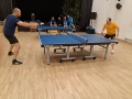 Dwaj mężczyźnia grają w tenisa stołowego, w tle siedzi czterech innych uczestników..