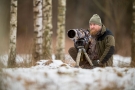 autor wystawy z aparatem na statywie w zimowej scenerii między drzewami