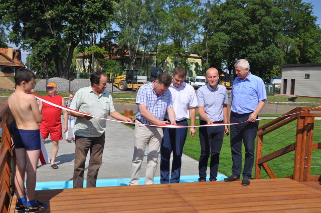 Na zdjęciu za szarfą stoi pięciu mężczyzn, jeden z nich ją przecina. Po lewej stronie Ratownik wodny ubrany w czerwony str&oacute;j i chłopiec w kapiel&oacute;wkach.