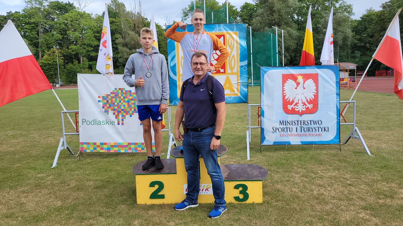 Zawodnicy na podium, przed stoi trener. W tle widać flagi, herby Ministerstwa Sportu i Turystyki oraz Województwa Podlaskiego 