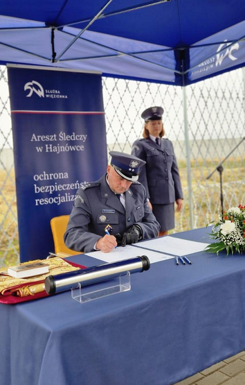 Przy stole siedzi funkcjonariusz podpisujący dokumenty. Z tyłu stoi funkcjonariuszka.