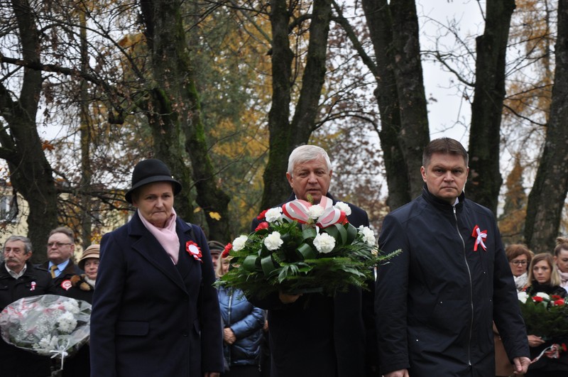 delegacja trzech osób .Męzczyzna w środku niesie wieniec z biało-czerwonych kwiatów. 