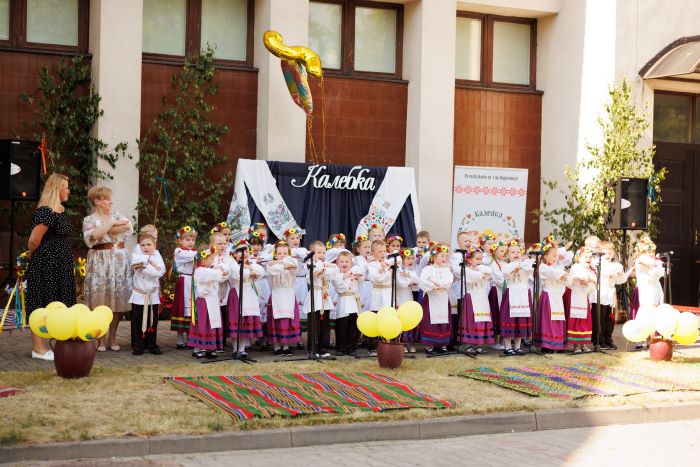 Dzieci z grupy Kolebka ubrane w ludowe stroje śpiewają przed mikrofonami, obok stoją dwie nauczycielki.