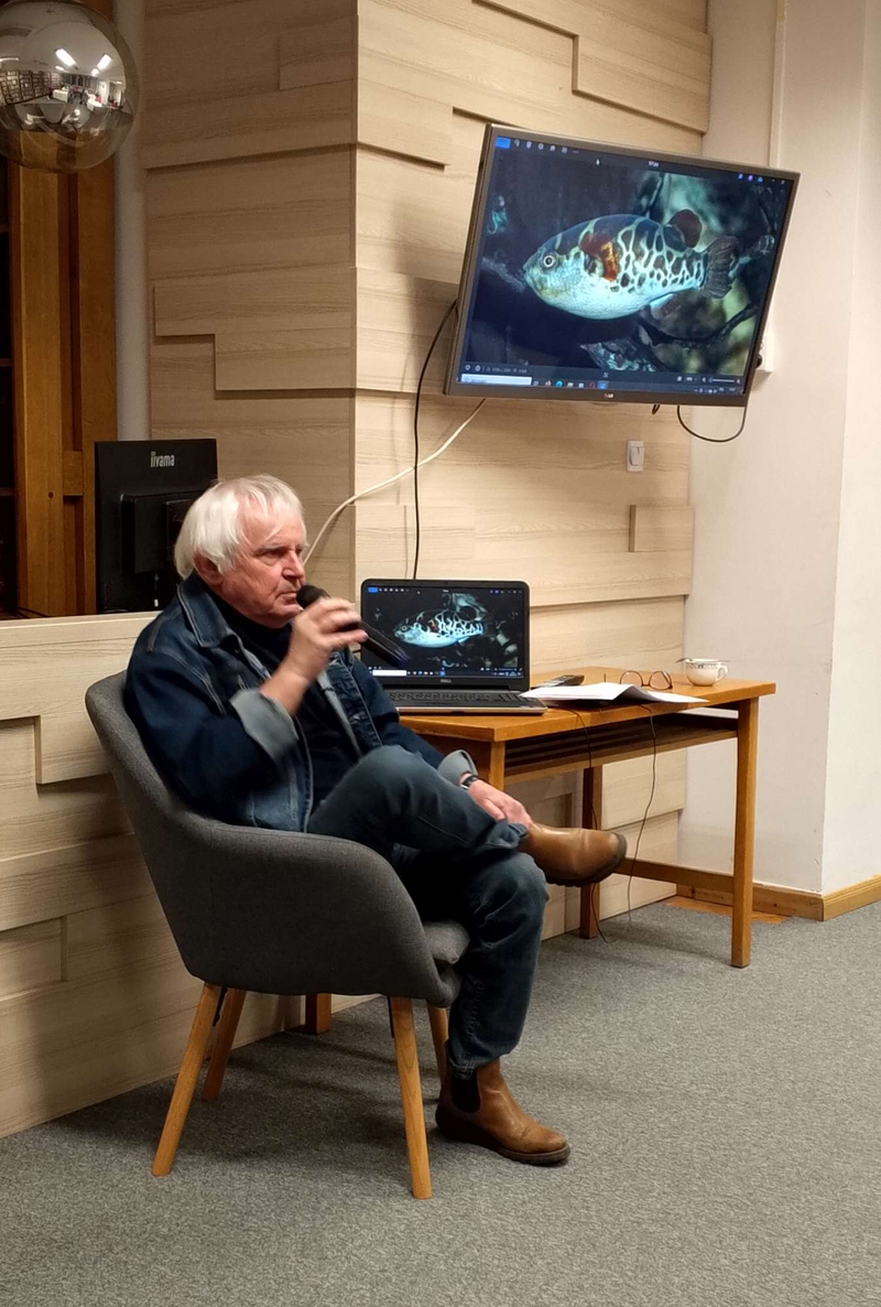 mężczyzna siedzi na fotelu, w tle widać zdjecie ryby wyświetlane na telewizorze