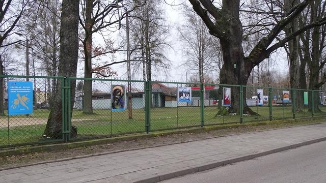 prace konkursowe zawiezone na ogrodzeniu; za nim rosną wysokie drzewa