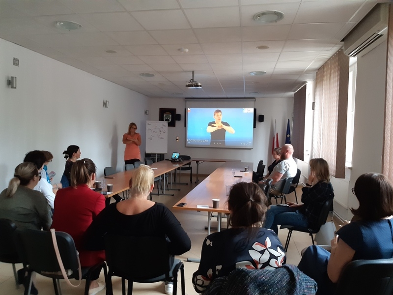 sala konferencyjna: przy stołach siedzą uczestnicy szkolenia, przed nimi prowadzący, na ścianie wisi ekran z wyświetlonym slajdem