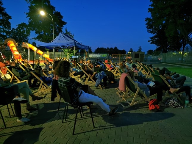 duża liczba widzów kina plenerowego siedąca na krzesłach i leżakach rozstawionych na placu. Na zewnętrz panuje już półmrok. Z tyłu widać element placu zabaw oraz rozstawiony namiot. Wokół rosną drzewa. Na tle jednego z nich znajduje się świecąca lampa uliczna