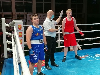 W ringu stoi dwóch zawodników, jeden z nich ubrany w czerwony, a drugi w niebieski strój bokserski. Pomiędzy nimi stoi sędzia z maseczką na twarzy.