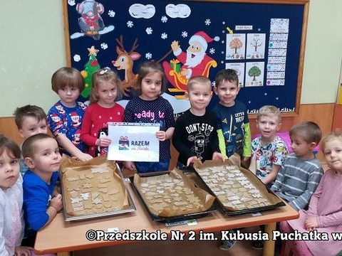 Dzieci siedzą wokół stolika z upieczonymi pierniczkami. Dwie dziewczynki trzymają kartkę z napisem "Razem na święta".