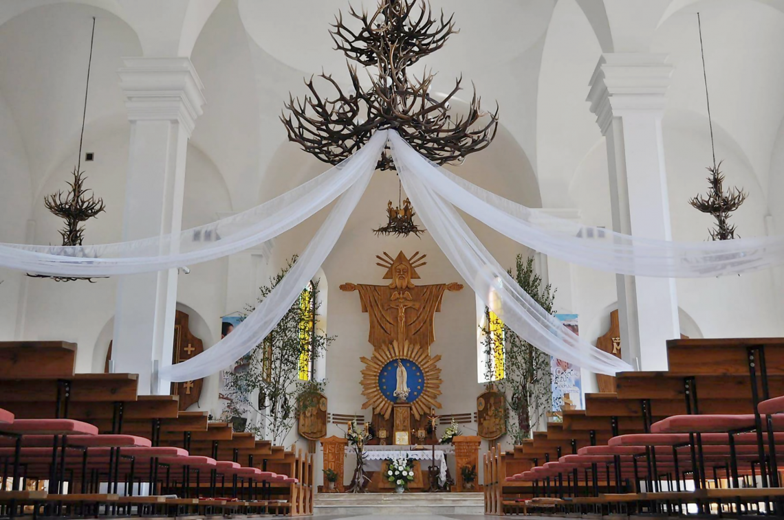 wnętrze świątyni w którym przeważa biel; po prawej i lewej stronie ustawione są drewniane ławki; na wprost widnieje ołtarz wykonany z drewna; z góry zwisają żyrandole z poroży jeleni